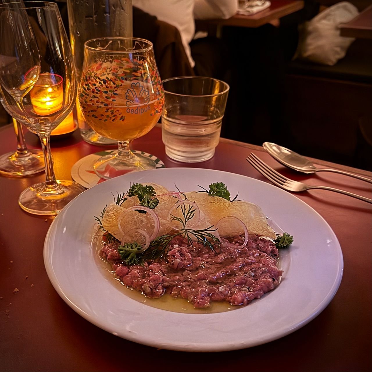 Beef tartare topped with crispy chips and fresh dill on a white plate. A full glass of beer stands within reach beside it.