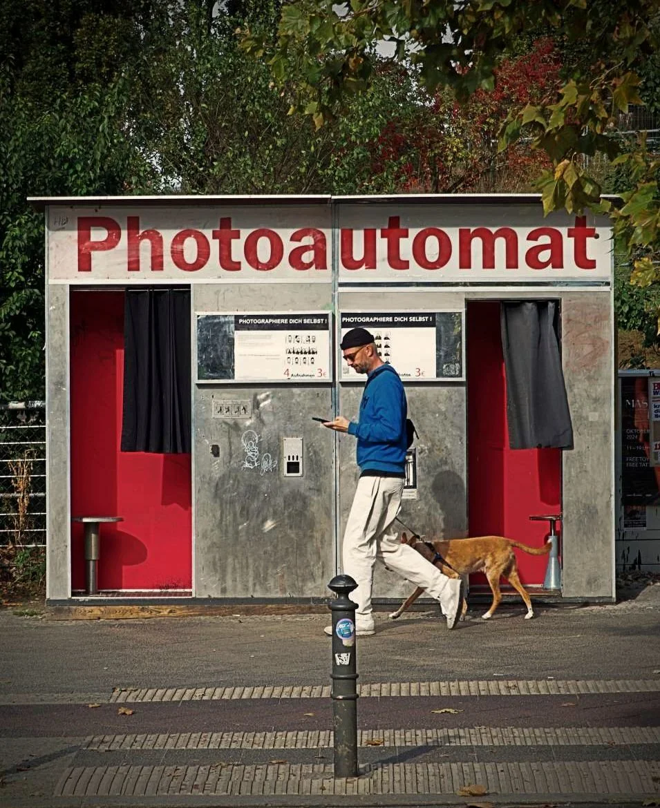 Historischer Photoautomat in Berlin mit roter Beschriftung, zwei Kabinen und Vorhang. Davor geht ein Mann mit Hund vorbei, Asphaltstraße, Poller und herbstliche Bäume im Hintergrund.