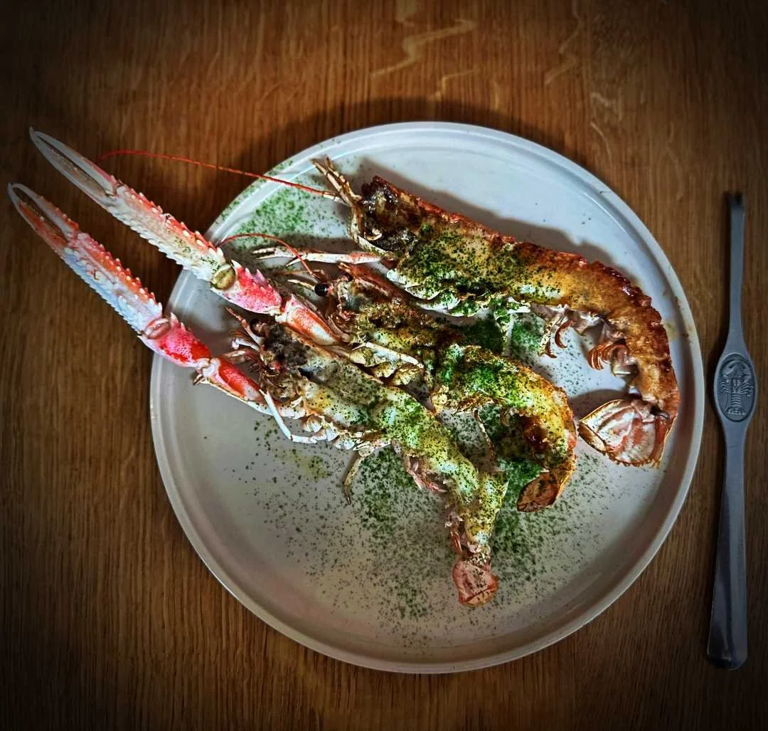 Langoustines from Iluka restaurant in Copenhagen, served on a light ceramic plate with grilled langoustines, green herb powder and cutlery on a wooden table.
