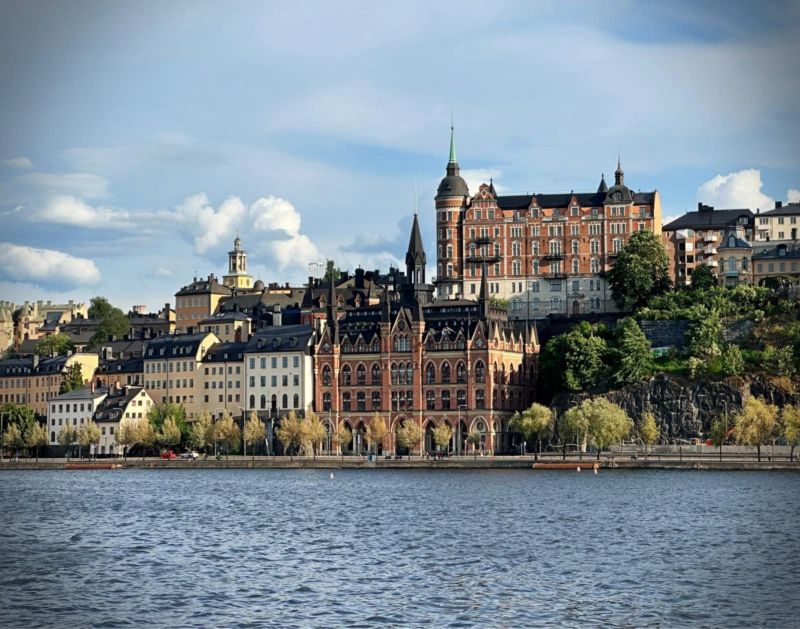 Blick über das Wasser auf Södermalm in Stockholm. Historische Backsteinbauten, helle Wohnhäuser und begrünte Felsformationen entlang des Ufers unter blauem Himmel.