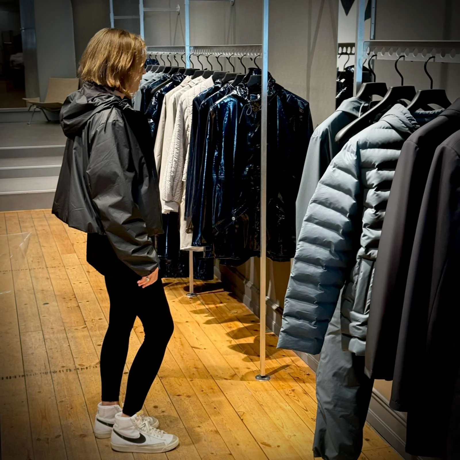 Eine Frau betrachtet Jacken an einem Kleiderständer im Rains Store, während sie eine kurze schwarze Regenjacke trägt.