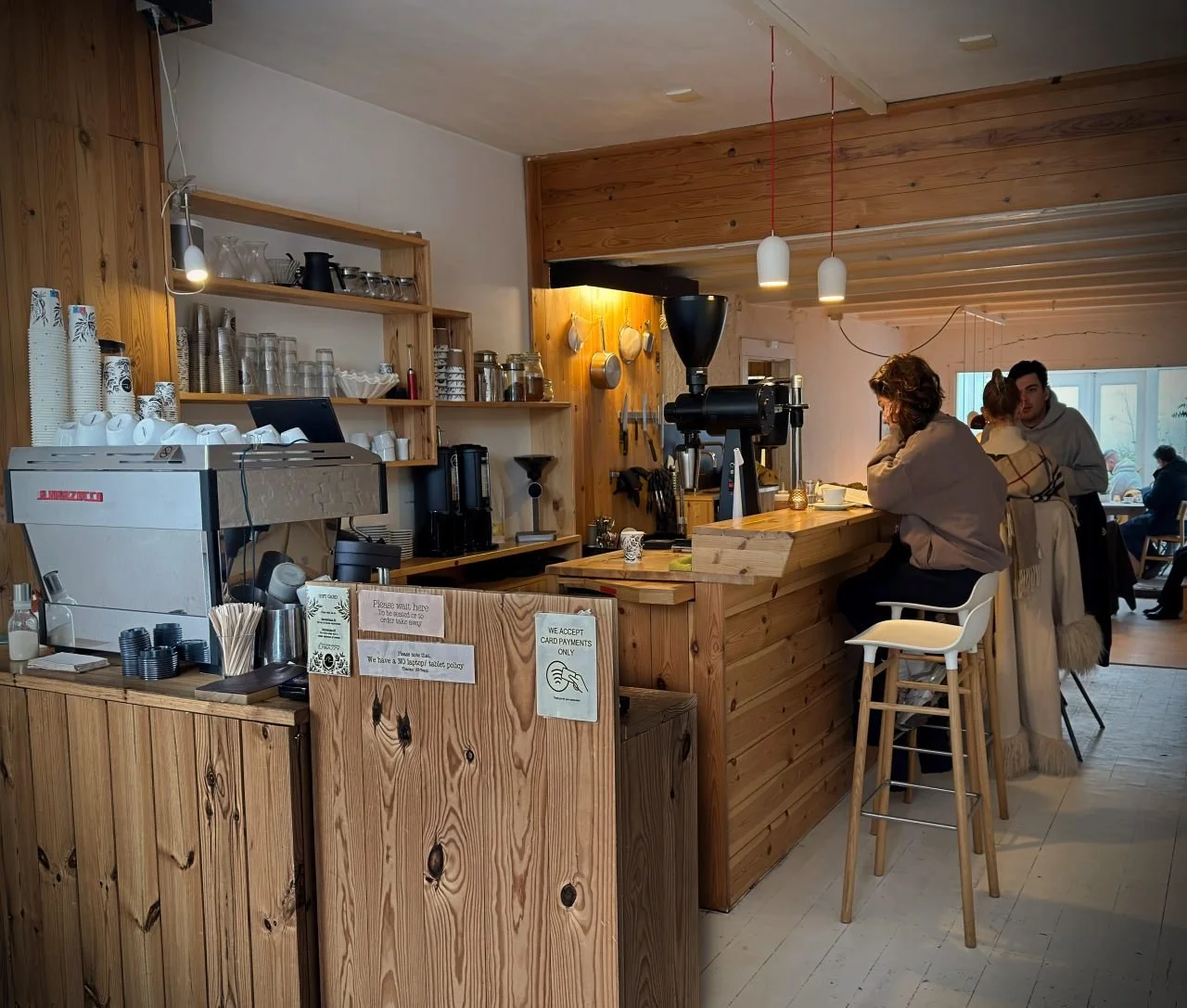 Interior of Scandinavian Embassy café in Amsterdam showing a wooden coffee bar, espresso machine, bar stools at the counter, and guests seated while waiting for their drinks.