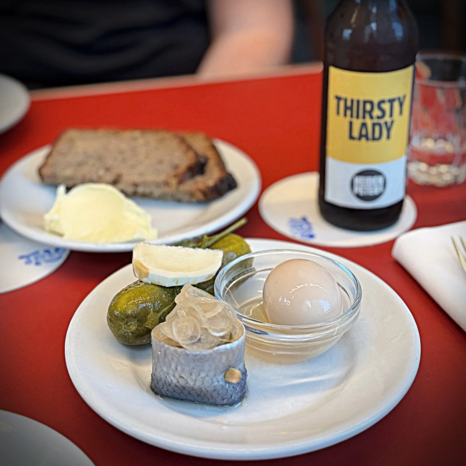 A plate with a rollmops, pickled cucumbers, a lemon wedge, and a pickled egg in a bowl on a red table. A plate with bread and butter and a bottle of beer stand behind it.