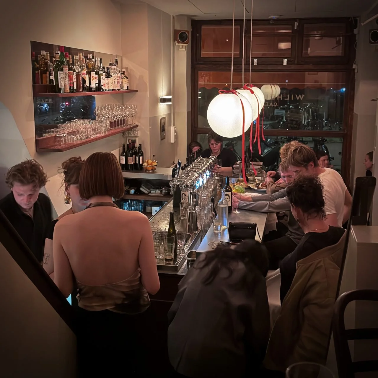 IView into the crowded taproom of Café Wilhelmina: Guests sit and stand close together at the long counter with its many beer taps, illuminated by large white spherical lamps.