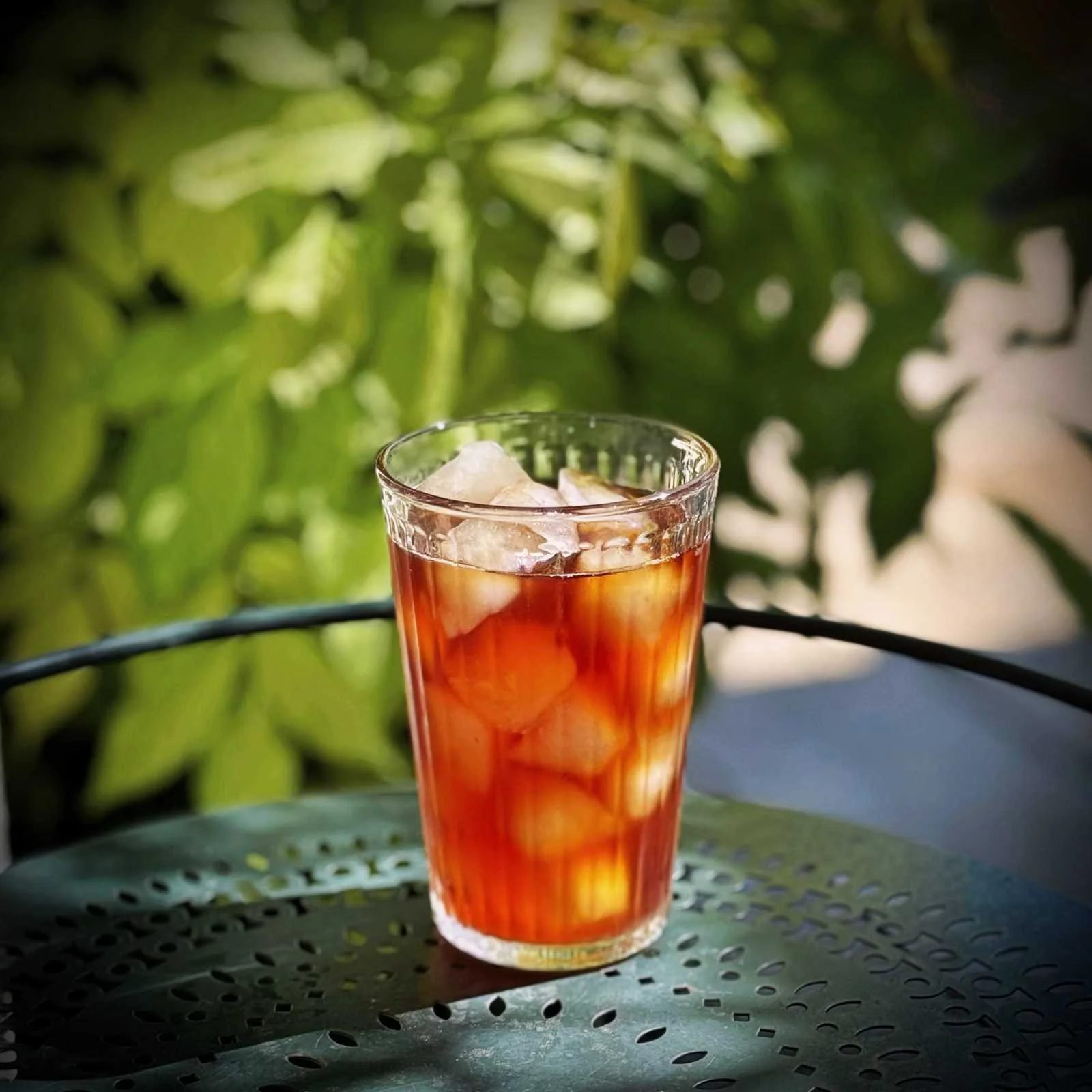 Ein transparentes Glas mit Eistee, Eiswürfeln, einer Zitronenscheibe und einem Strohhalm steht auf einer hölzernen Oberfläche. Im Hintergrund ist eine runde Metalldose erkennbar.