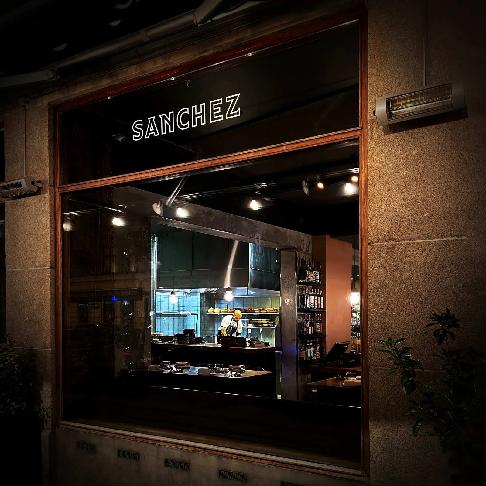 Blick durch das Schaufenster des Restaurants Sanchez in Kopenhagen Vesterbro auf die offene Küche und das Sanchez-Logo bei Nacht.