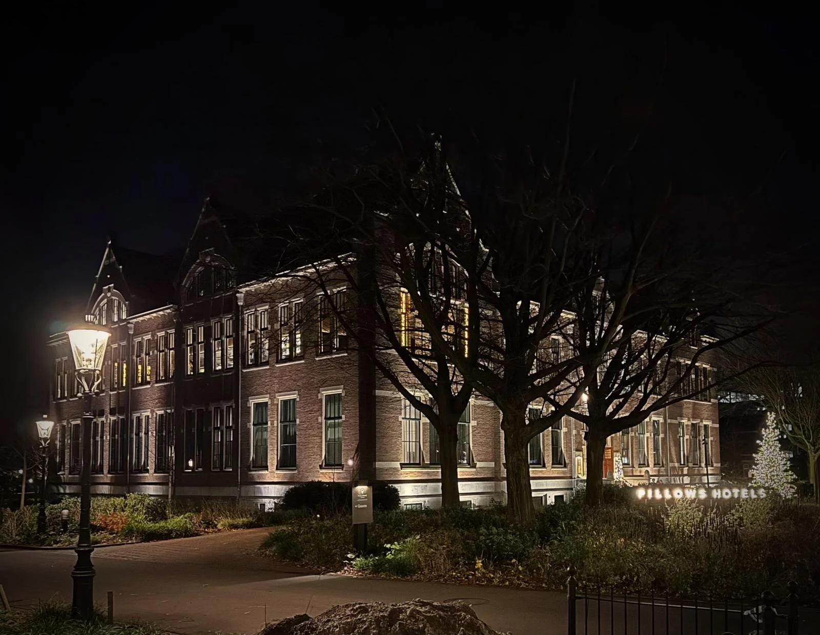 Nächtliche Backsteinfassade des Pillows Hotels Amsterdam, umgeben von Bäumen und traditionellen Laternen.