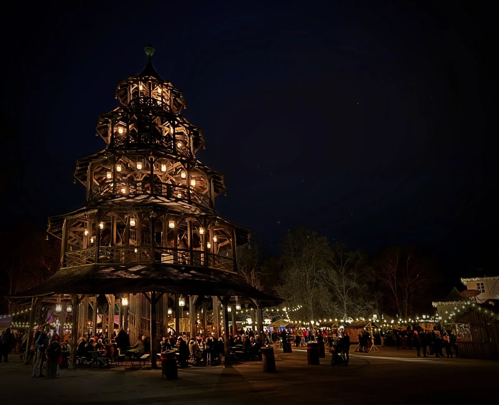 In the evening light the Chinese Tower in Munich’s English Garden is lit by lanterns, visitors sit on beer benches, and festive Christmas market stalls glow in the background.