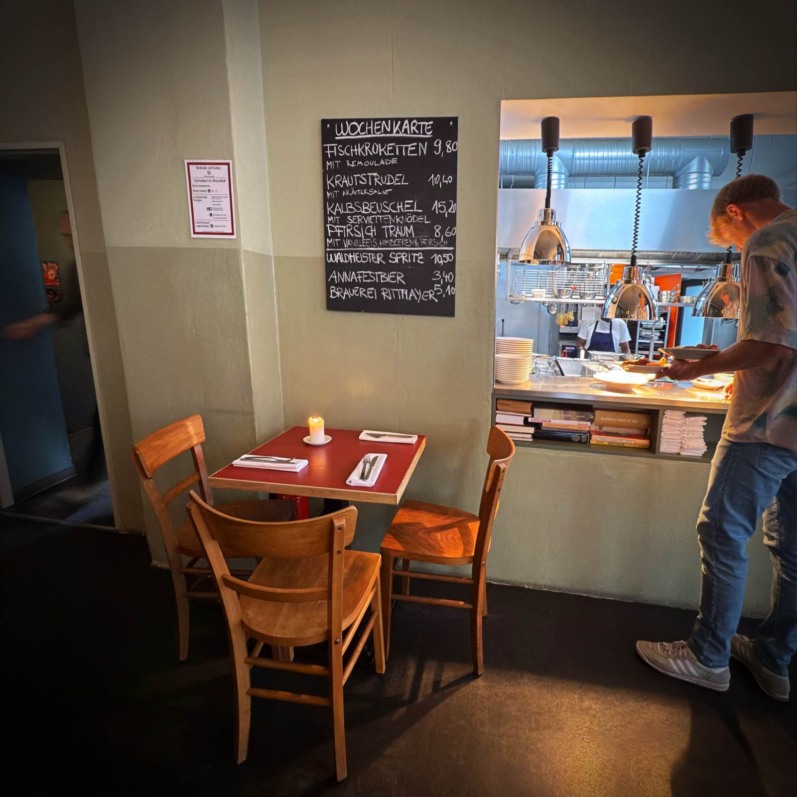 A small wooden table in front of a handwritten weekly menu board on the wall. On the right, a waiter stands at the kitchen pass picking up food.