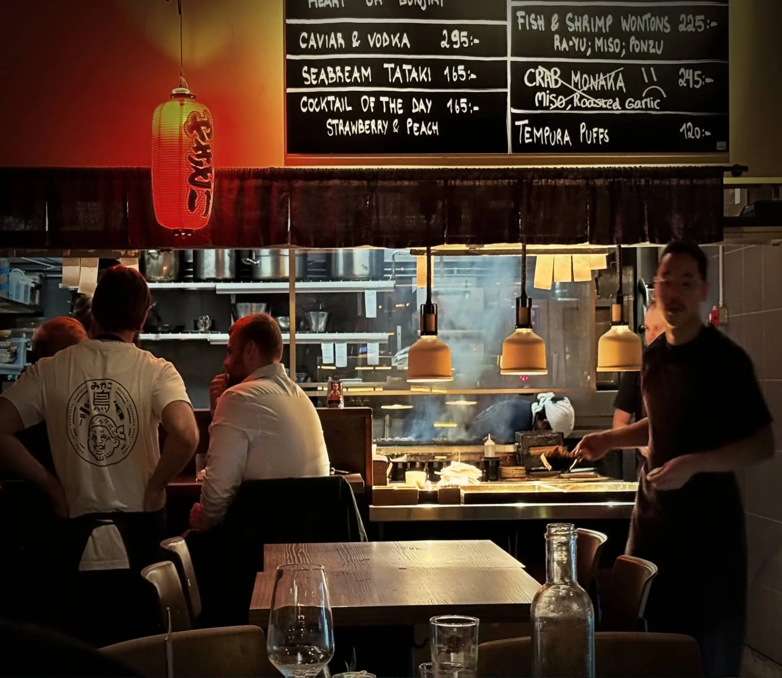 View of the open kitchen at Miyakodori restaurant in Stockholm featuring staff, menu boards, and a dining table in the foreground.