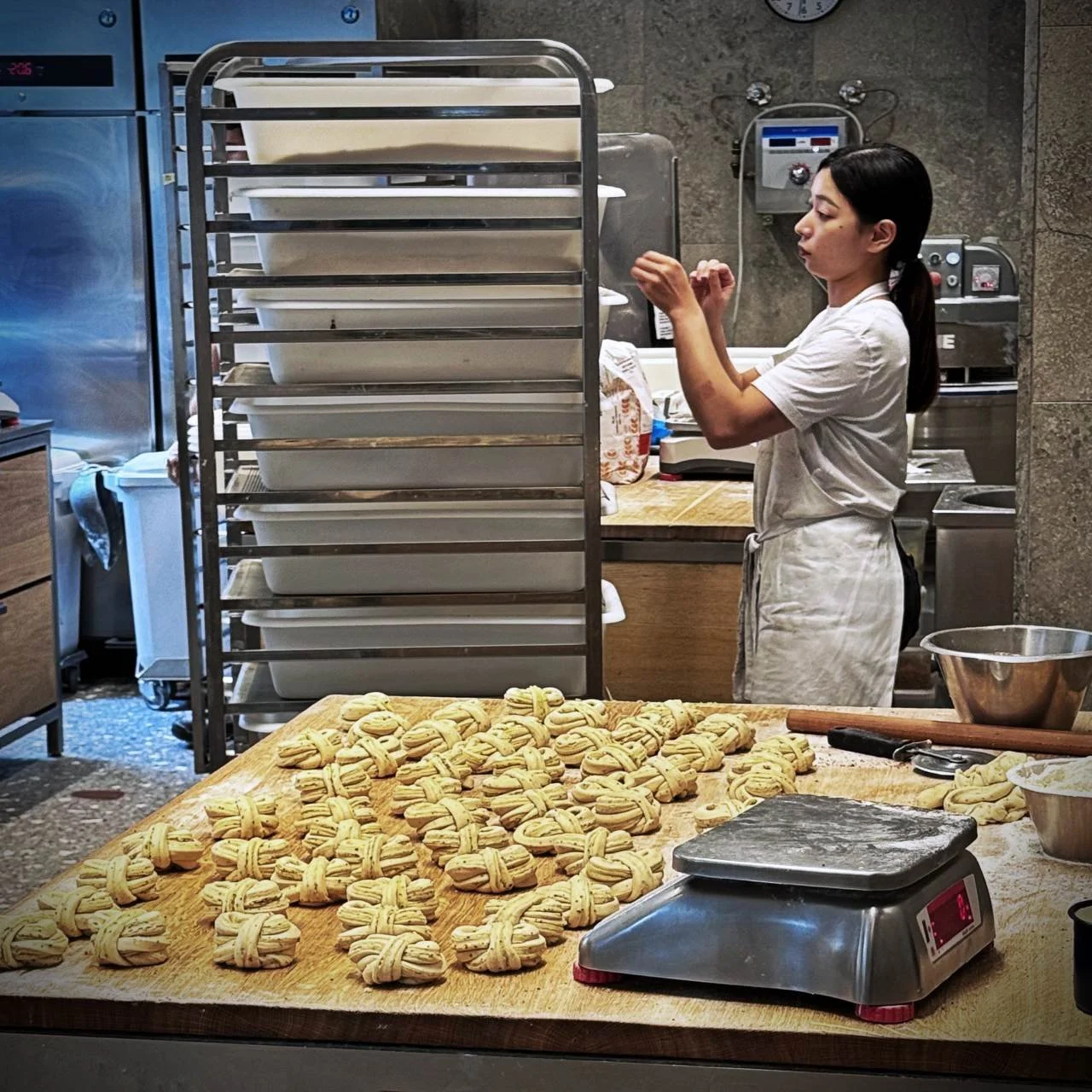In einer Backstube auf Holzplatten vorbereitete Zimtschnecken, im Hintergrund eine Mitarbeiterin in Weiß vor einem hohen Blechwagen mit Teigbehältern.