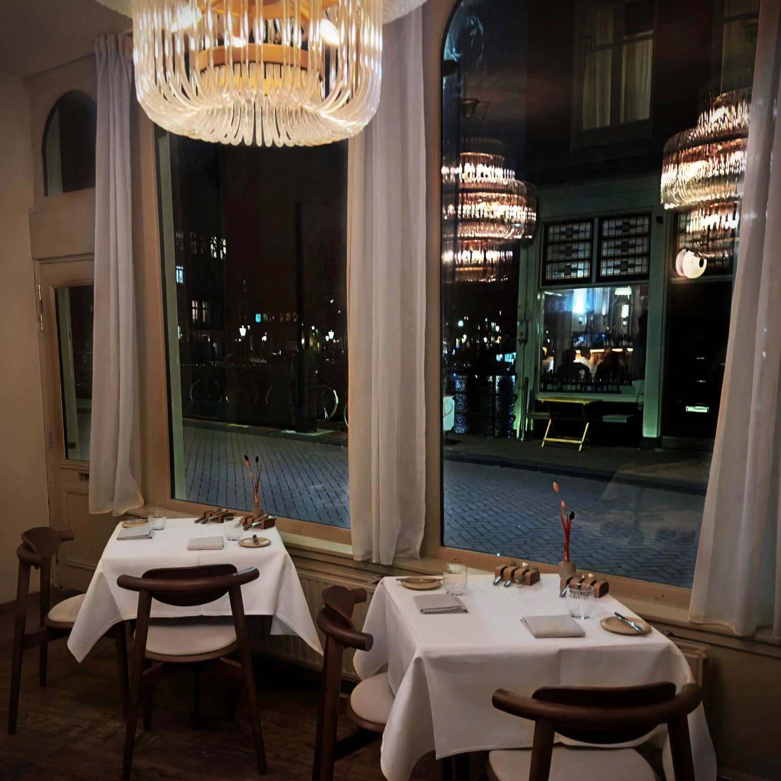 Im Restaurant R21 in Amsterdam stehen zwei eingedeckte Tische mit weißen Decken vor großen Fenstern. Ein großer Glaskronleuchter spiegelt sich nachts in der Scheibe über dem Parkettboden.