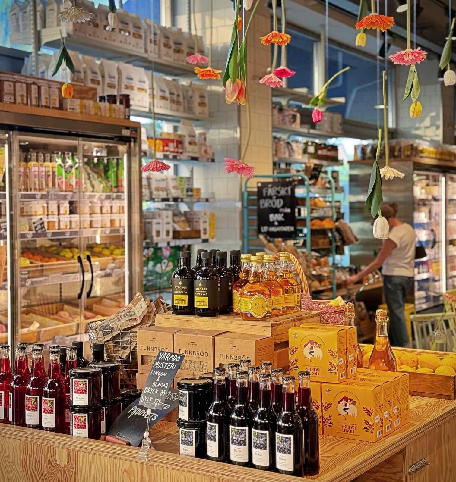 A retail display inside Urban Deli showcasing various Swedish gourmet products such as oils, juices, jams, and crispbread. Colorful upside-down tulips hang from the ceiling above.