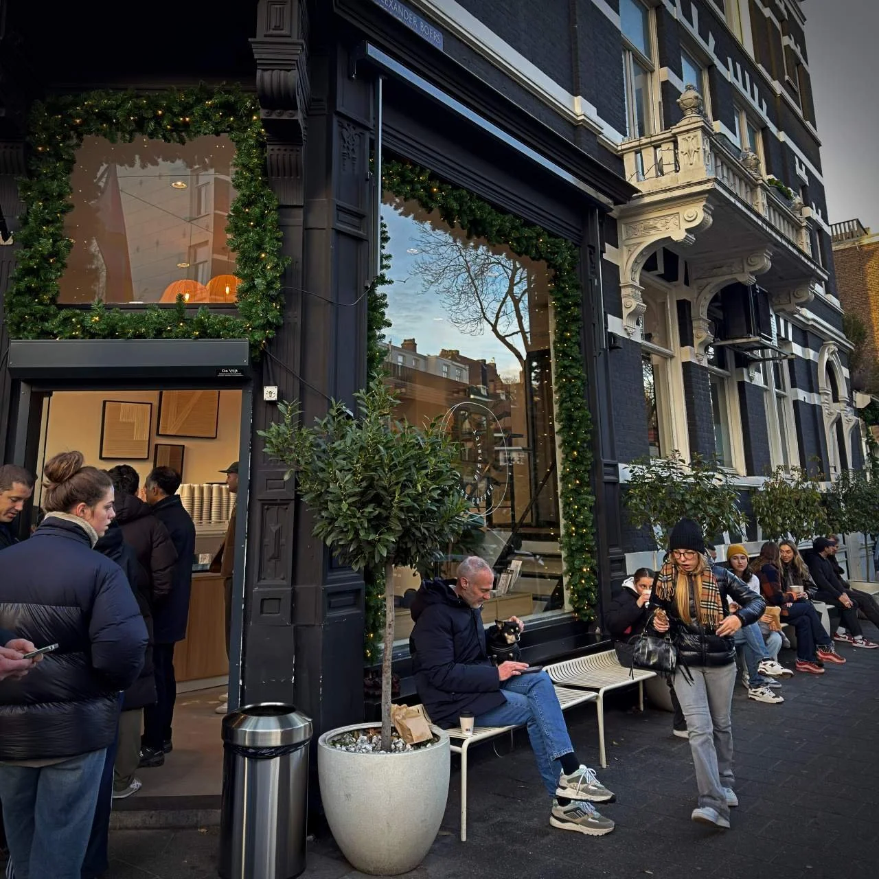 Gäste sitzen und stehen vor dem Coffee District am Willemsparkweg in Amsterdam, eingerahmt von grüner Deko und großen Fenstern im historischen Eckhaus.