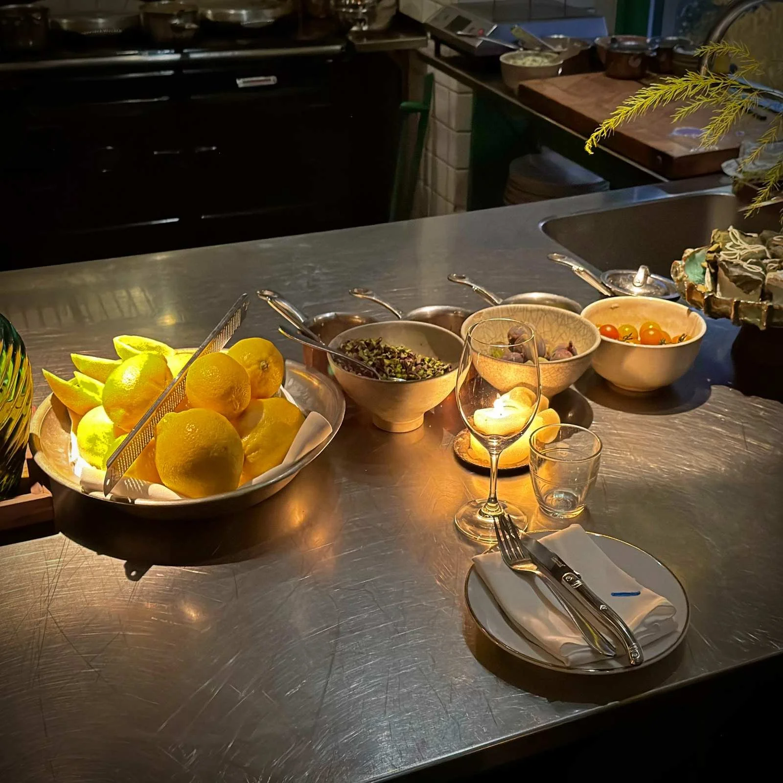 Eine Edelstahltheke mit einer Schale voller Zitronen und einer Reibe. Dahinter stehen mehrere Schüsseln mit Zutaten wie Pistazien, Feigen und Kirschtomaten sowie ein gedeckter Teller mit Besteck und Gläsern.