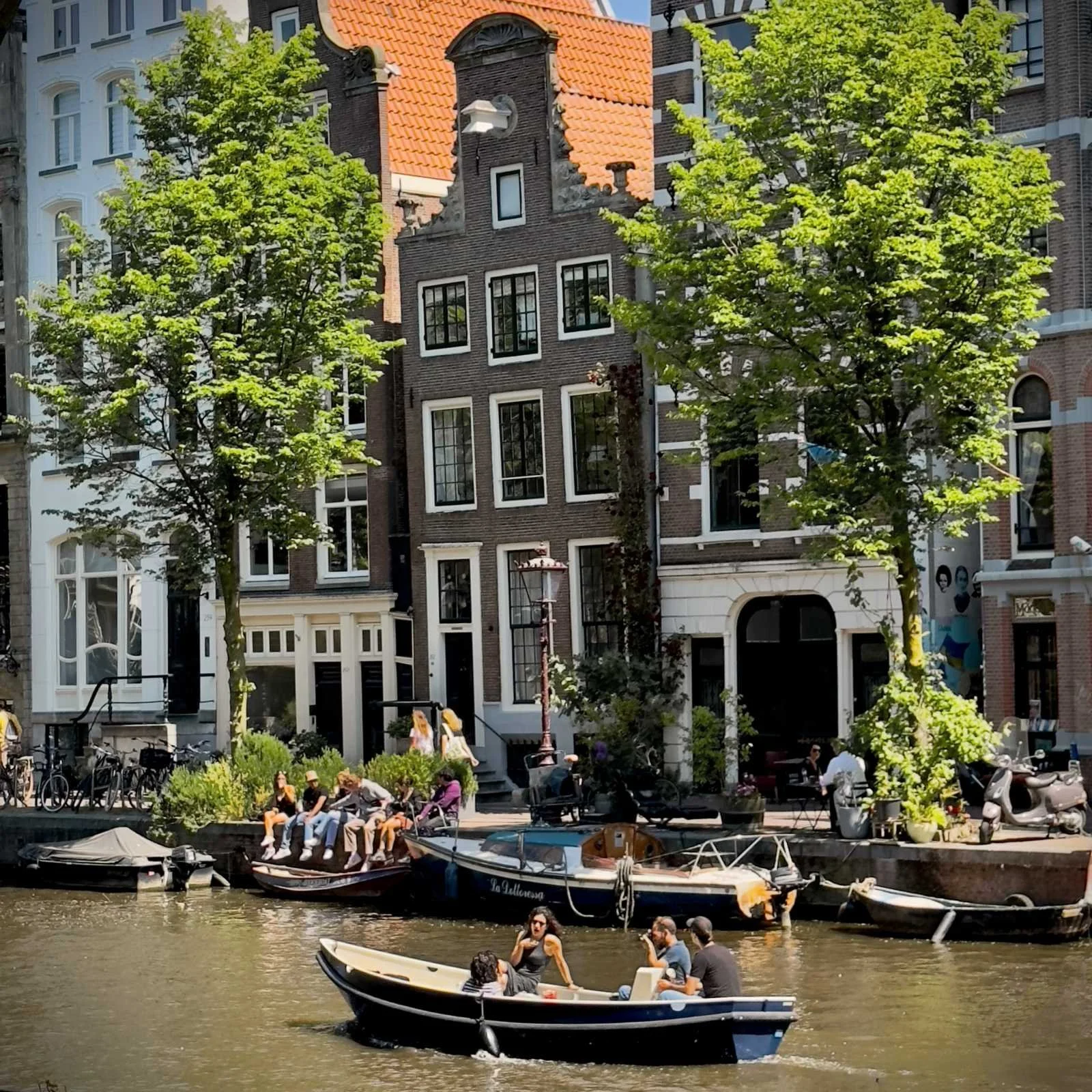A boat travels through a canal in the sun. People sit on the quay wall in front of the gabled houses.