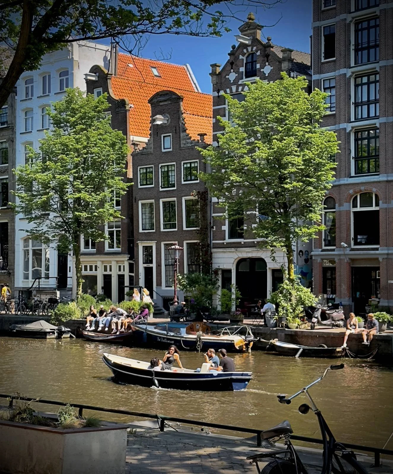 An open boat travels through the canal in the sunshine. People sit on the quay wall with their legs over the water, with narrow brick facades rising between the trees behind them.