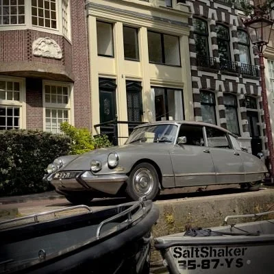 A grey vintage Citroën parked along a canal in front of narrow Amsterdam townhouses. The image represents the blend of heritage and urban lifestyle.