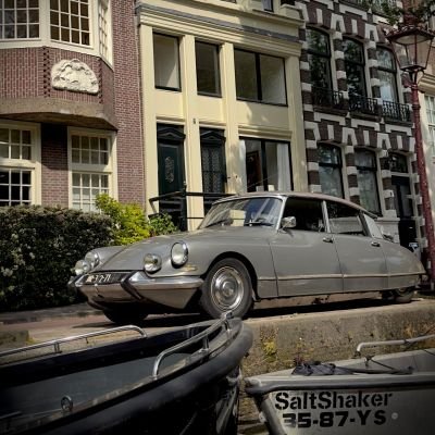 Ein klassischer Citroën DS parkt am Ufer einer Gracht vor historischen Amsterdamer Giebelhäusern. Das Bild fängt die nostalgische Atmosphäre und den urbanen Charme der niederländischen Hauptstadt ein.