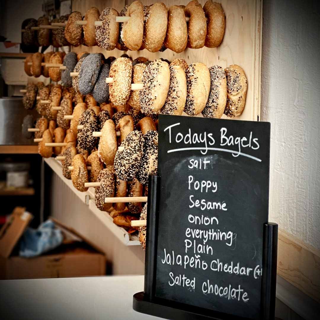 Verschiedene Bagels im Breadwinner Café in Amsterdam, auf Holzstäben präsentiert. Ein Schild zeigt Sorten wie Sesam, Jalapeño Cheddar und Salz-Schokolade.