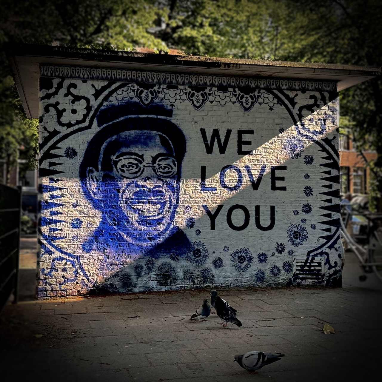 A blue portrait of the singer André Hazes is painted on a brick wall. Next to it are the words "WE LOVE YOU". Two pigeons sit on the ground in the foreground.