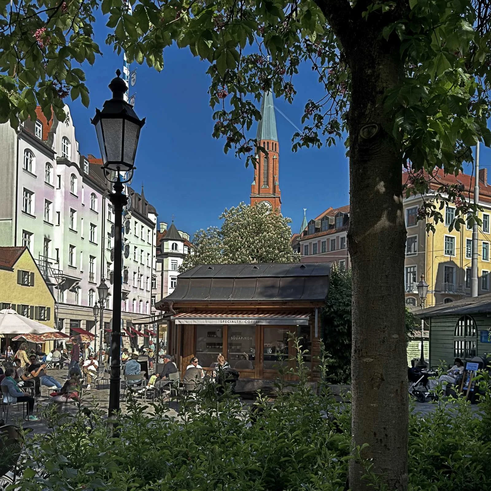 People sitting at tables in front of the Suuapinga coffee stall at Wiener Platz in Munich. The view includes a classic street lamp, trees, and a church tower against a blue sky in the background.