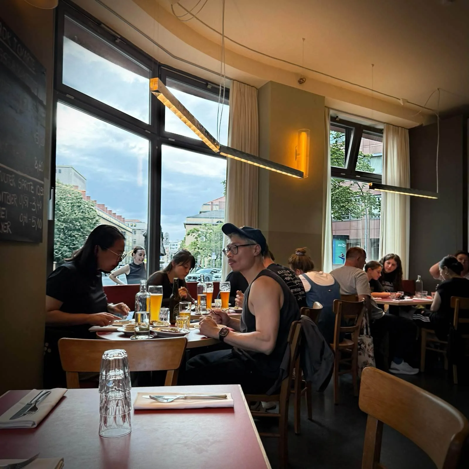 The dining area of Restaurant Trio with guests seated at tables in front of large window fronts. Elongated light fixtures hang from the ceiling, and a labeled board hangs on the left wall.