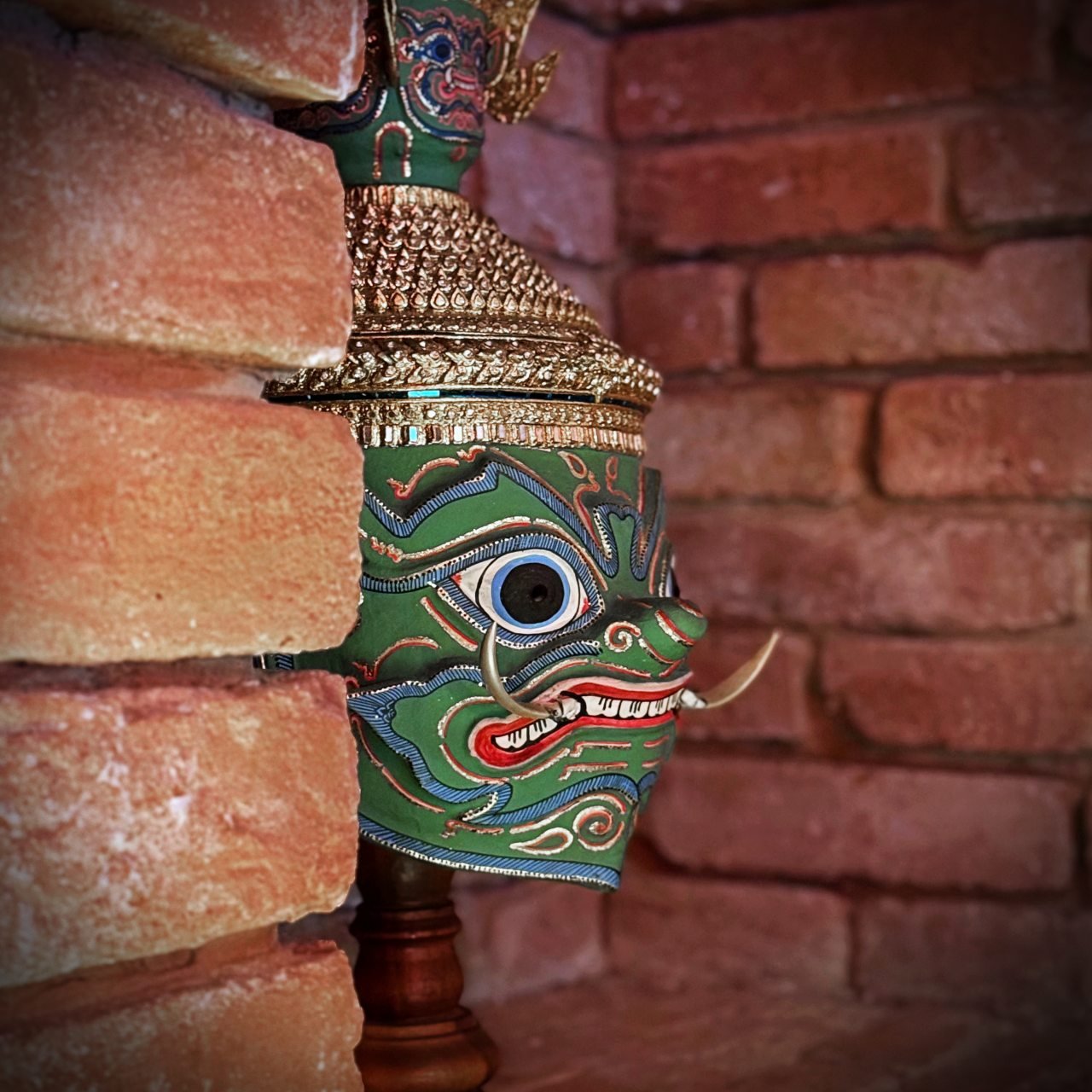 Detailaufnahme einer thailändischen Maske mit grünem Gesicht, goldener Kopfbedeckung und Stoßzähnen, inszeniert vor einer Wand aus roten Ziegelsteinen.