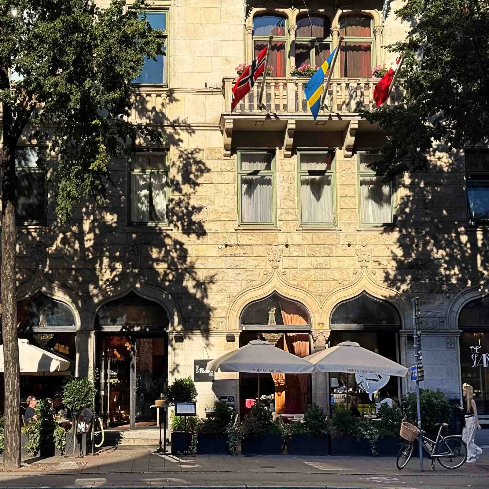 Außenansicht der Natursteinfassade von The Sparrow Hotel in Stockholm. Am Balkon im oberen Stockwerk sind drei Flaggen montiert.Im Erdgeschoss befindet sich der Außenbereich des Hotelrestaurants