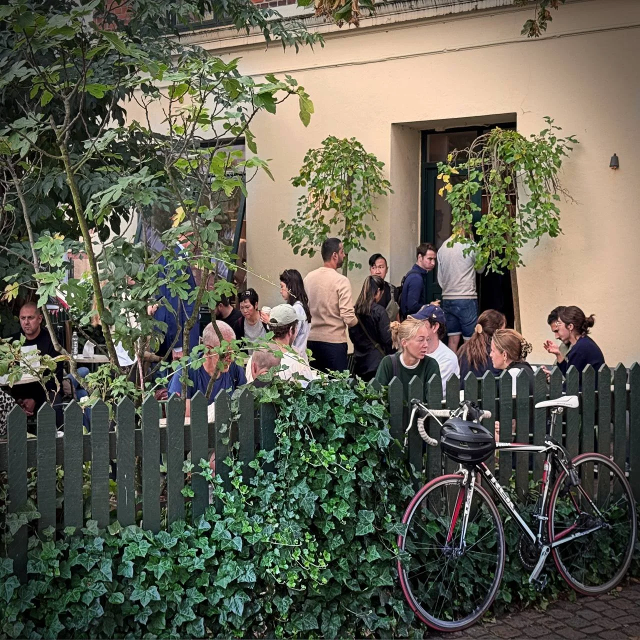Gäste stehen im Garten von Juno the Bakery in einer Schlange bis vor die Tür. Andere sitzen bereits an Tischen hinter dem mit Efeu bewachsenen Zaun, an dem ein Rennrad lehnt.