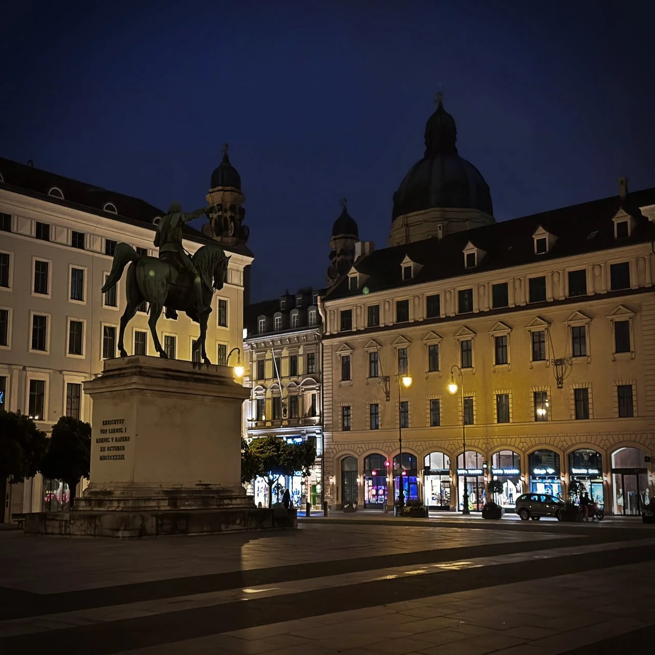 Reiterstatue von Ludwig I. auf dem nächtlich beleuchteten Wittelsbacherplatz in München, umgeben von historischen Fassaden und geöffneten Ladengeschäften.