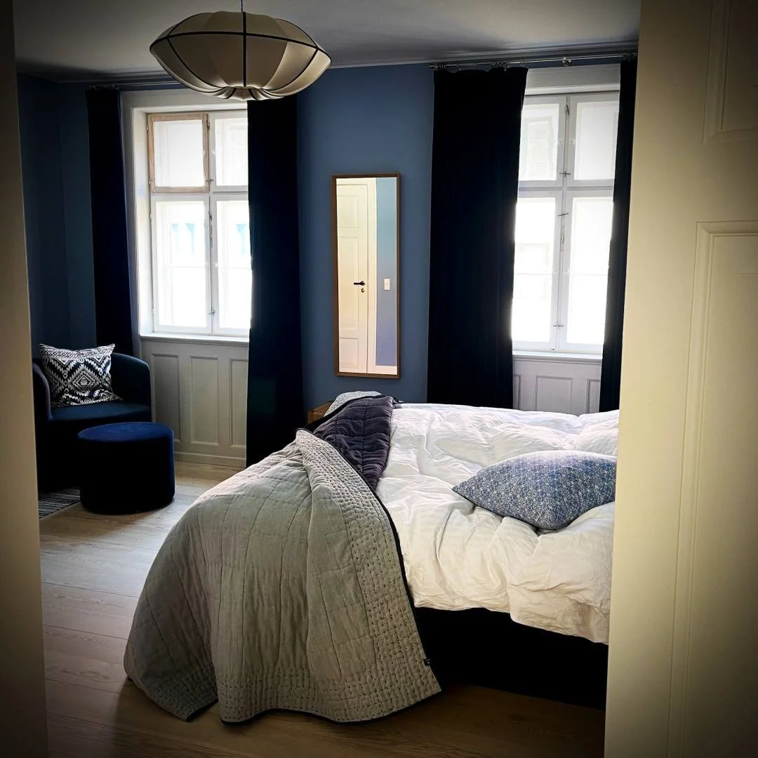 View into the bedroom of an Airbnb on Haregade in Copenhagen. The room has blue painted walls and is furnished with a double bed featuring a grey quilt, a blue armchair with a pouf, and a wall mirror between two windows.