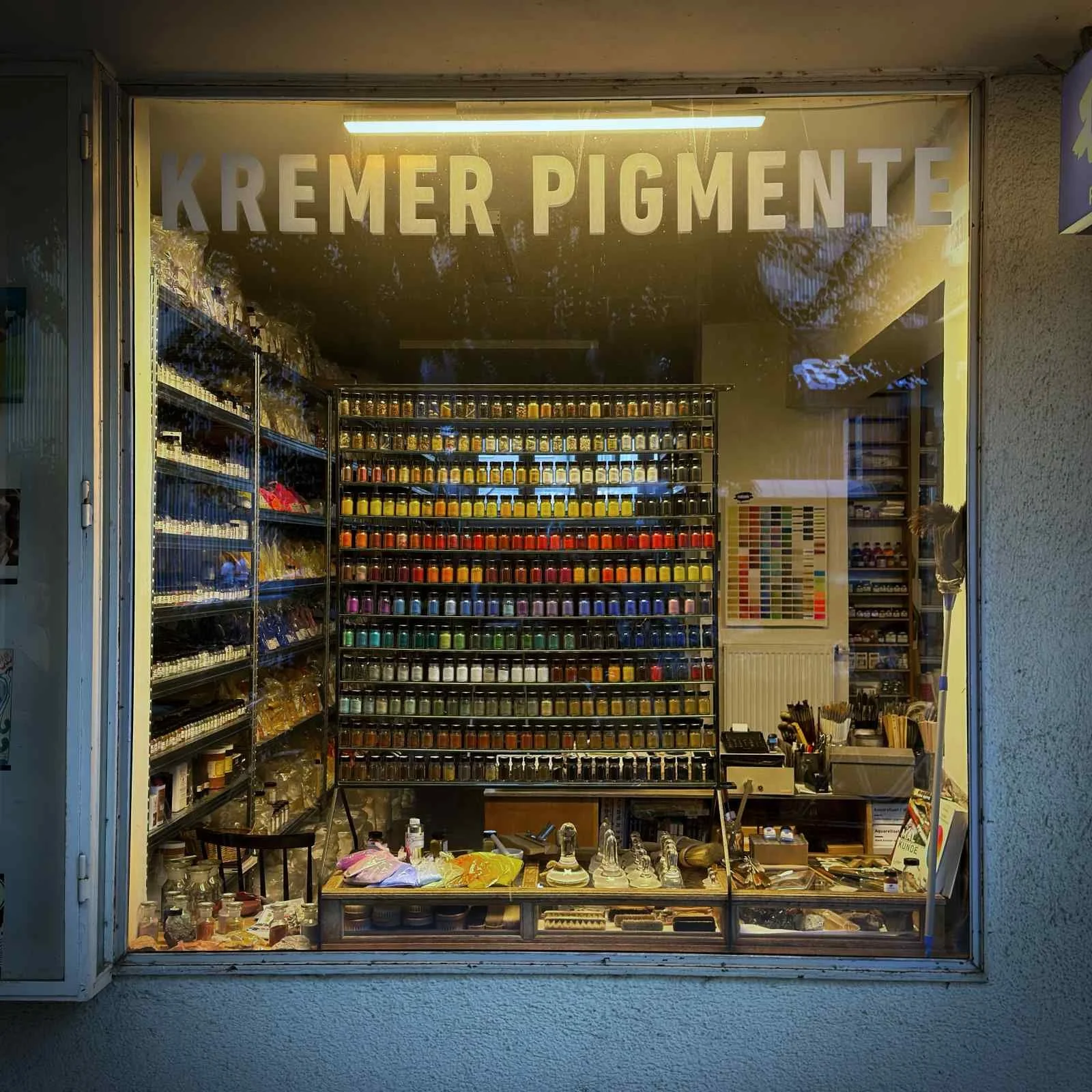 Schaufenster des Ladens Kremer Pigmente mit einer farblich sortierten Wand aus zahllosen Gläsern voller Farbpigmente.