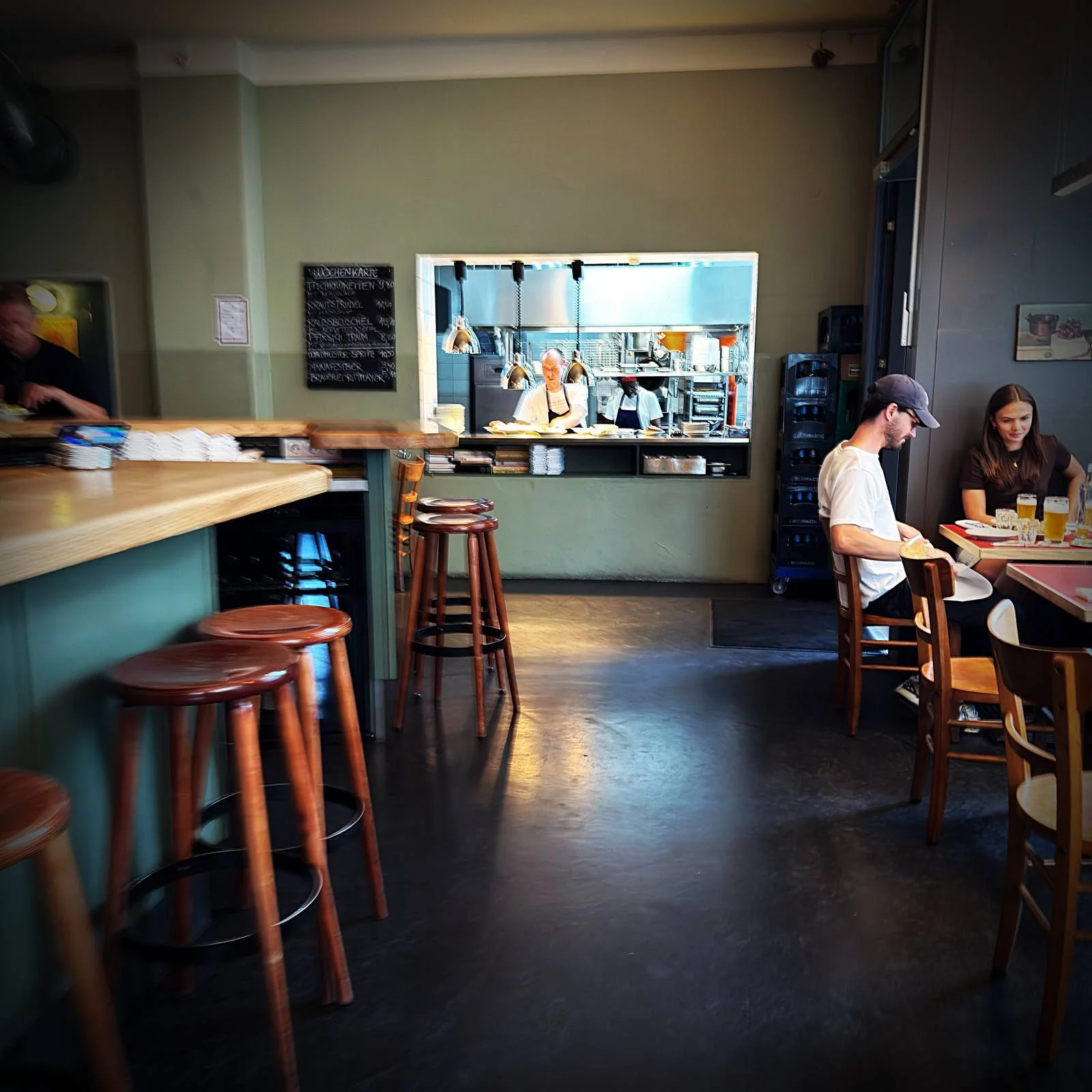 Interior view of Restaurant Trio with wooden stools at the bar in the foreground. The kitchen pass is visible in the background, with guests seated at tables on the right.