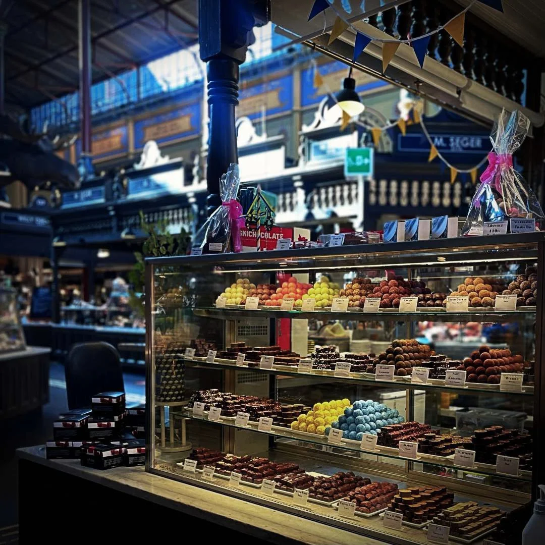 Eine gläserne Vitrine in der Östermalms Saluhall ist prall gefüllt mit handgemachten Pralinen und bunten Trüffeln. Im Hintergrund erkennt man die historische Holzarchitektur der Markthalle und dekorative blau-gelbe Wimpel.