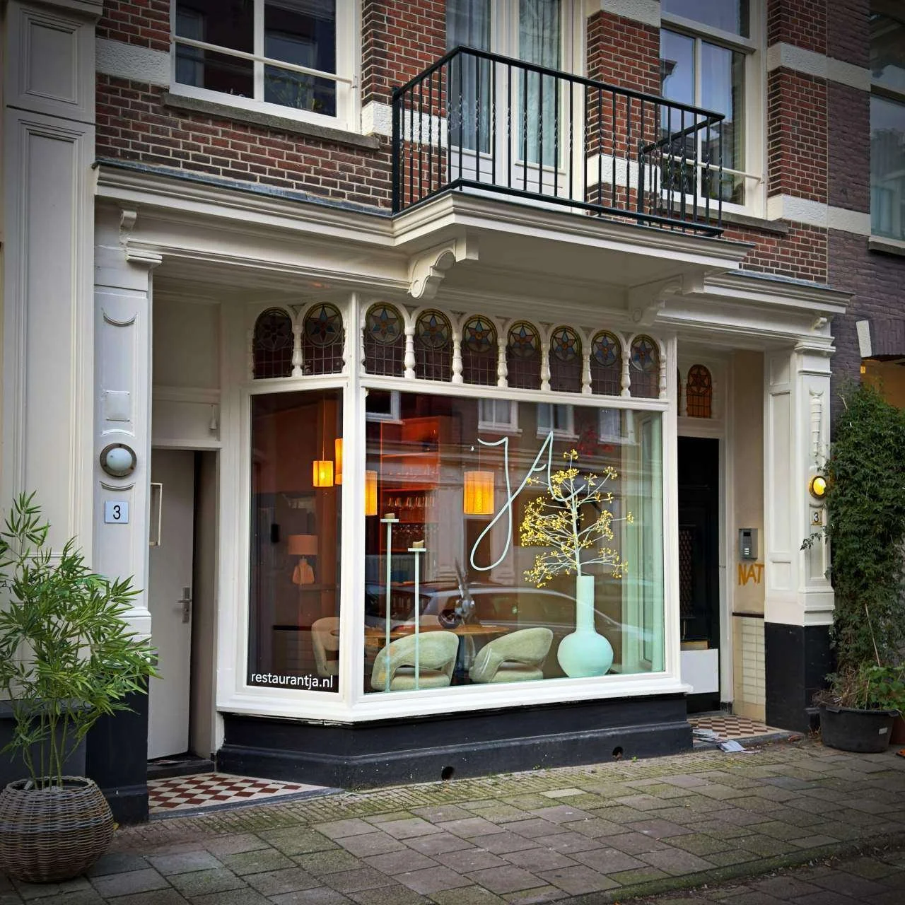 Exterior view of Restaurant Jā in Amsterdam with large front window, floral vase display, Art Nouveau details and website printed on the glass.