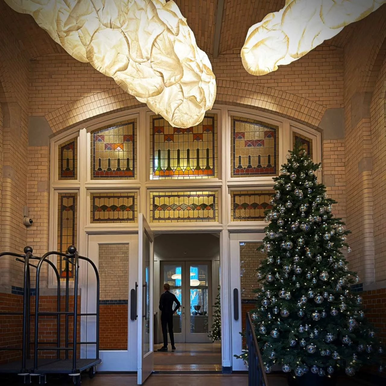 Festliche Eingangshalle im Pillows Hotel Amsterdam mit Weihnachtsbaum, historischen Bleiglasfenstern und modernen Wolkenlampen.