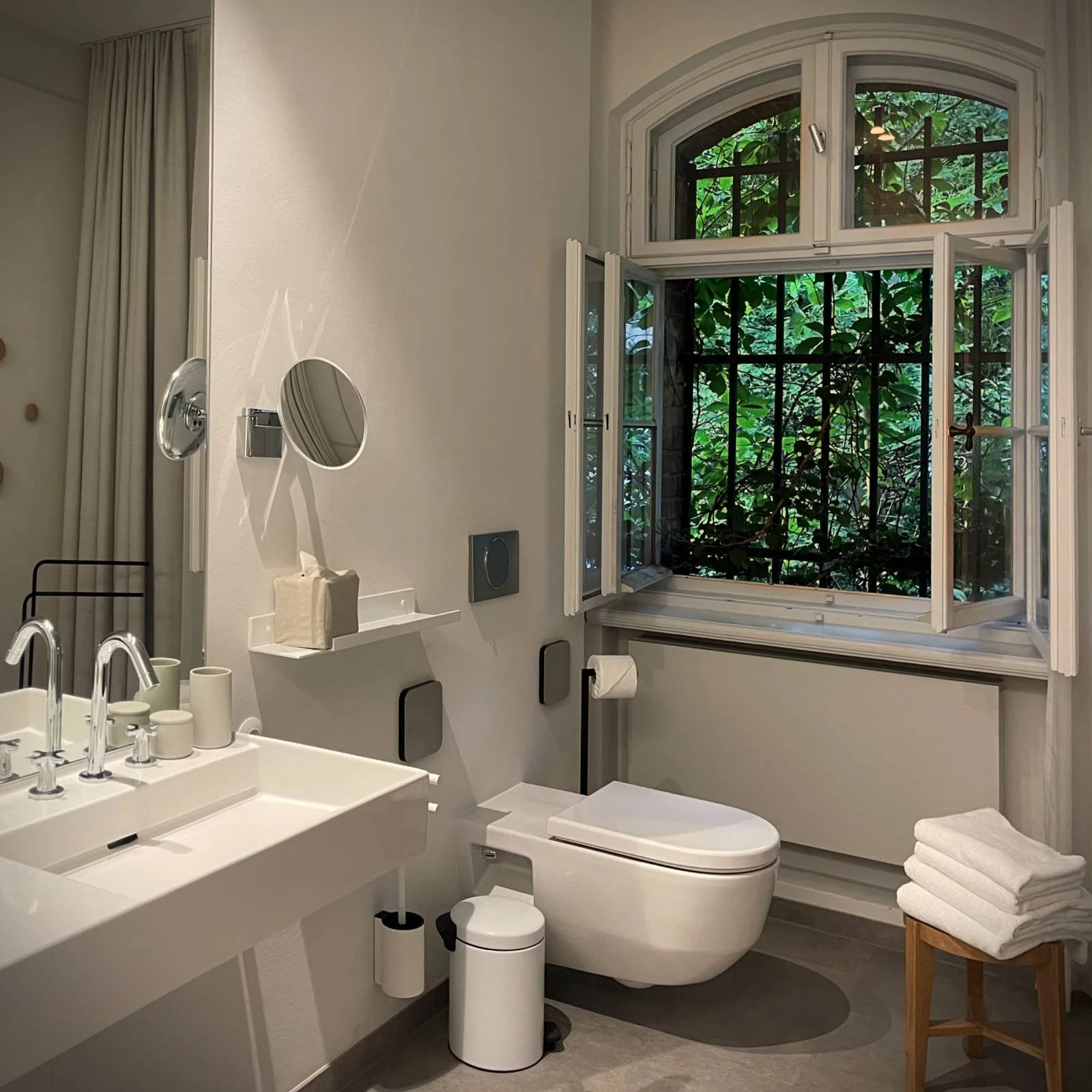 Ein Badezimmer im Berliner Hotel Wilmina mit einem offenen Fenster und Blick in den grünen Garten. Zur Ausstattung gehören ein weißes Waschbecken, ein Wandspiegel, eine Toilette und weiße Handtücher auf einem Holzhocker.