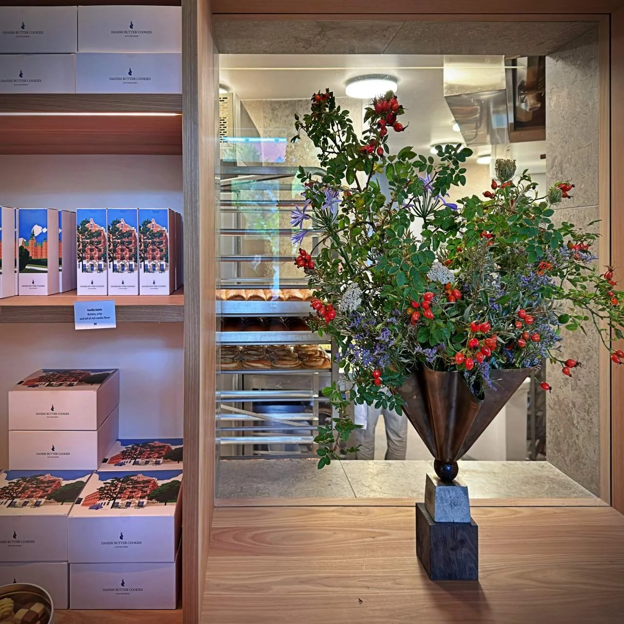 Blick auf Verkaufsregal mit Gebäckboxen, daneben ein großer Strauß mit Wildblumen, durch das Fenster ist das Backblechregal sichtbar.