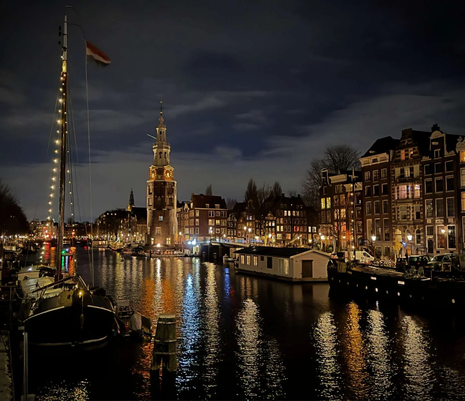 Eine nächtliche Ansicht des Montelbaanstoren in Amsterdam am Kanal. Boote liegen am Ufer, während sich die Lichter der umliegenden Häuser und des Turms im dunklen Wasser spiegeln.