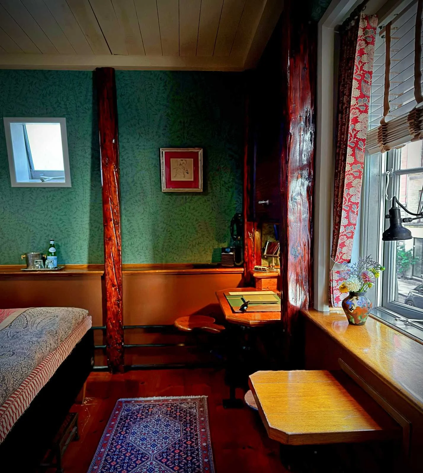 Gemütliches Hotelzimmer mit grüner Mustertapete, dunklen Holzelementen, einem kleinen Schreibtisch und einem Fenster mit Blick nach draußen.