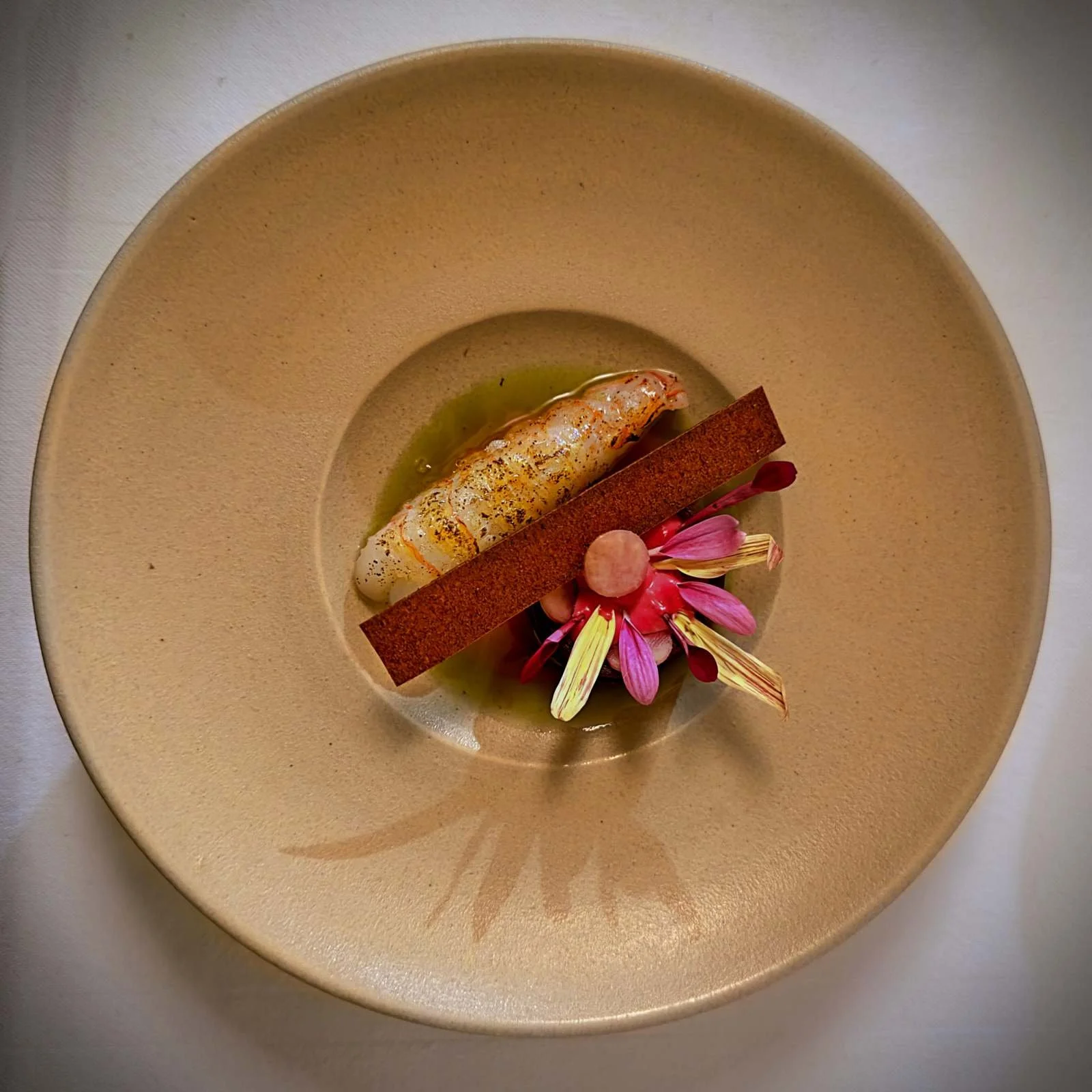 Auf einem beigen Teller ist eine Langustine mit Roter Beete und Feige angerichtet. Ein brauner, knuspriger Riegel und rosa Blütenblätter ergänzen die Vorspeise im Restaurant R21 in Amsterdam.