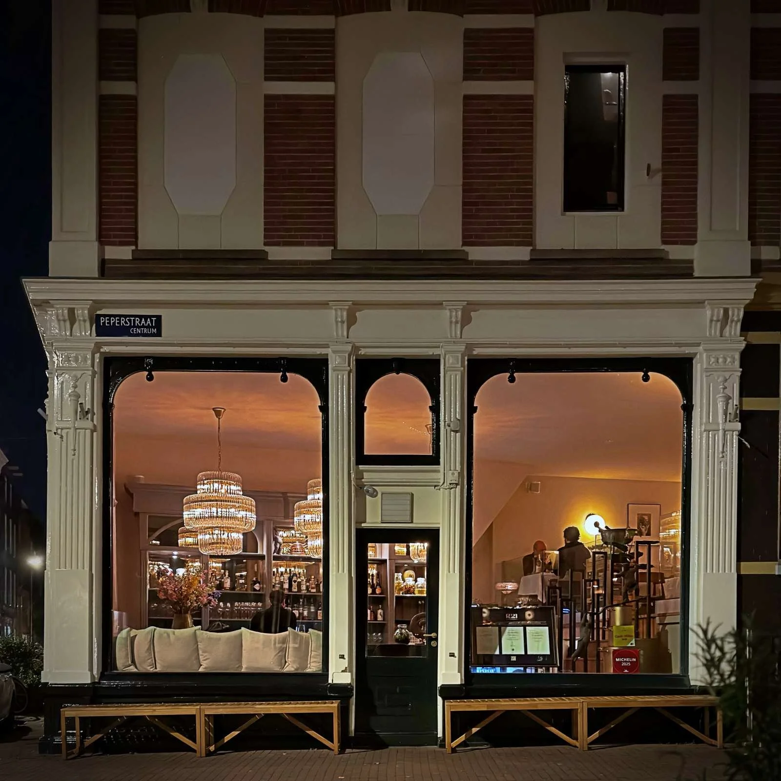 Die nächtliche Fassade des Restaurants R21 in der Peperstraat in Amsterdam zeigt zwei große Fenster. Im beleuchteten Innenraum sind Kronleuchter sowie ein Michelin-Aufkleber am Fenster erkennbar.