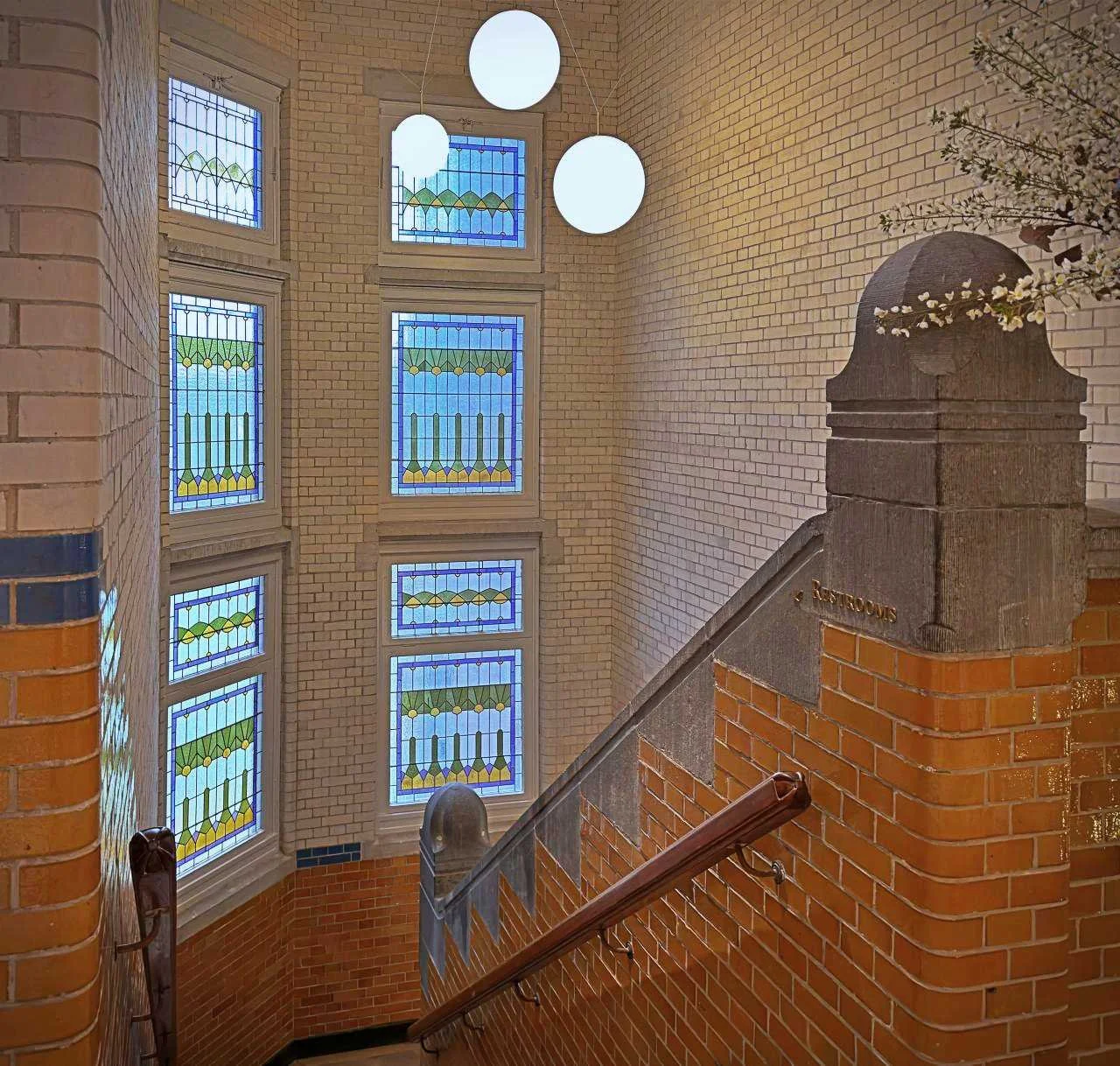 Historisches Treppenhaus im Pillows Hotel Amsterdam mit farbigen Bleiglasfenstern, Ziegelwänden und modernen runden Pendelleuchten.