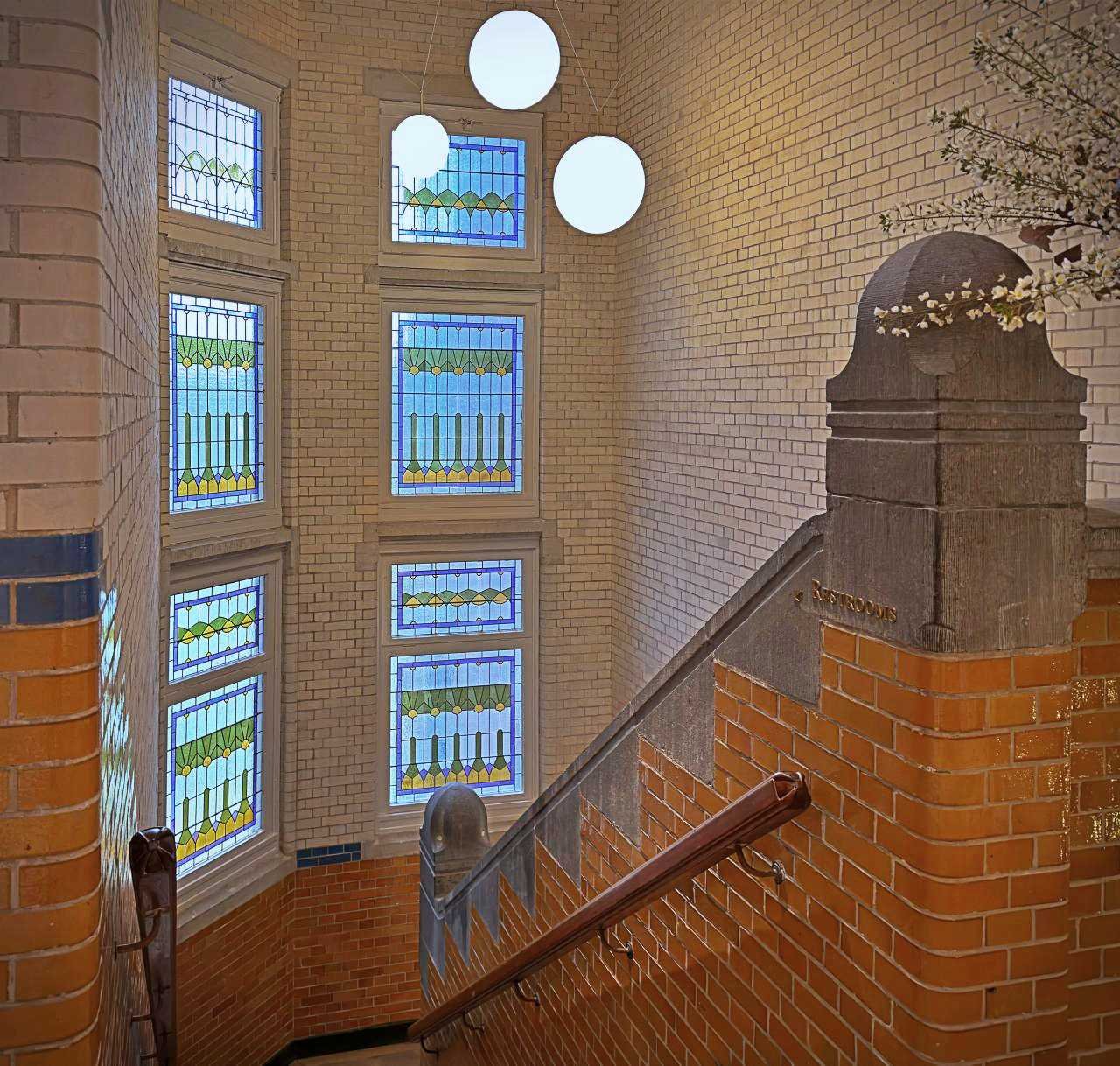 Historic staircase at Pillows Hotel Amsterdam featuring colorful stained glass windows, brick walls, and modern round pendant lights.