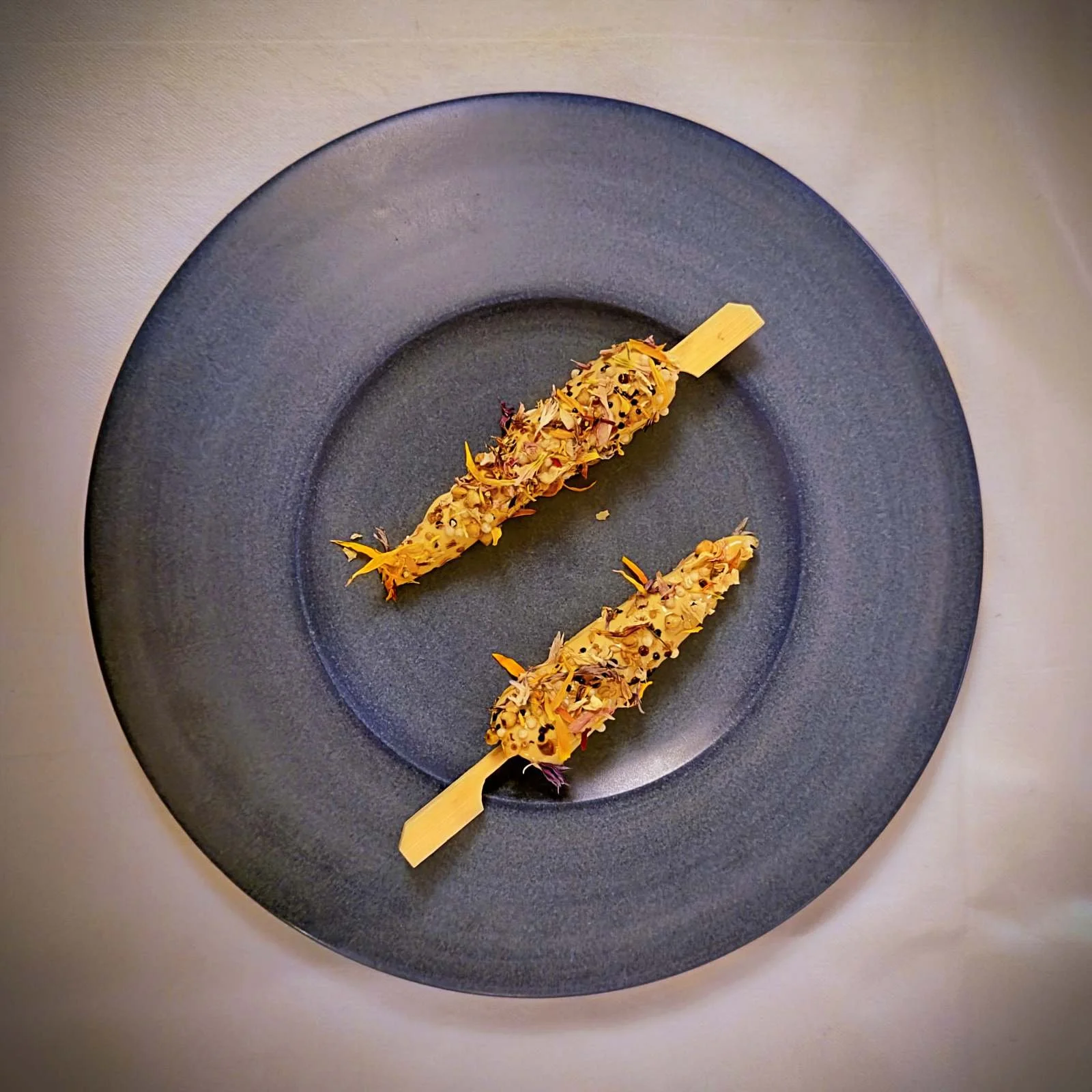 Auf einem dunkelblauen Teller liegen zwei Holzspieße mit Wolfsbarsch-Ceviche. Die Vorspeise im Restaurant R21 in Amsterdam ist mit gepufftem Mais und bunten Blütenblättern garniert.