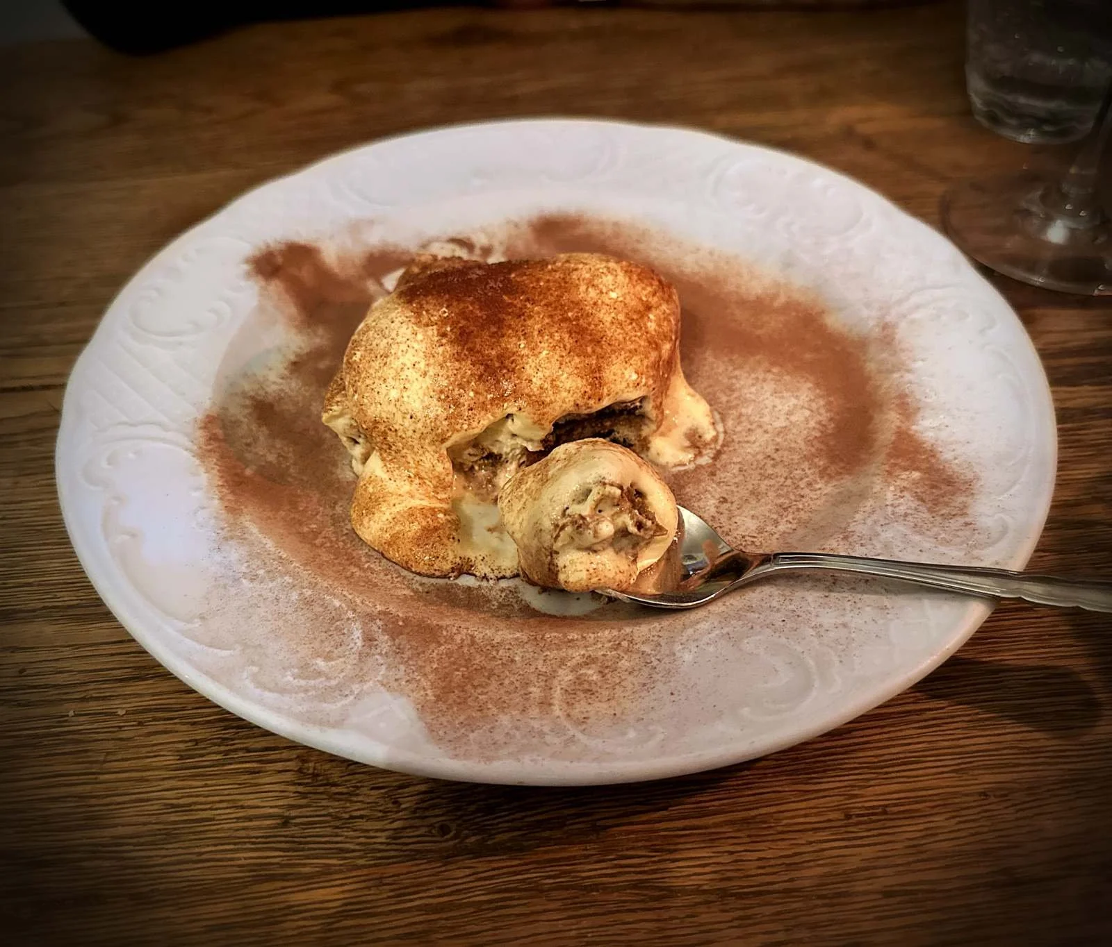 Ein Tiramisu auf einem weißen, mit Kakaopulver bestreuten Teller mit einem Löffel auf einem Holztisch im Restaurant Il Piccolo Principe in München.