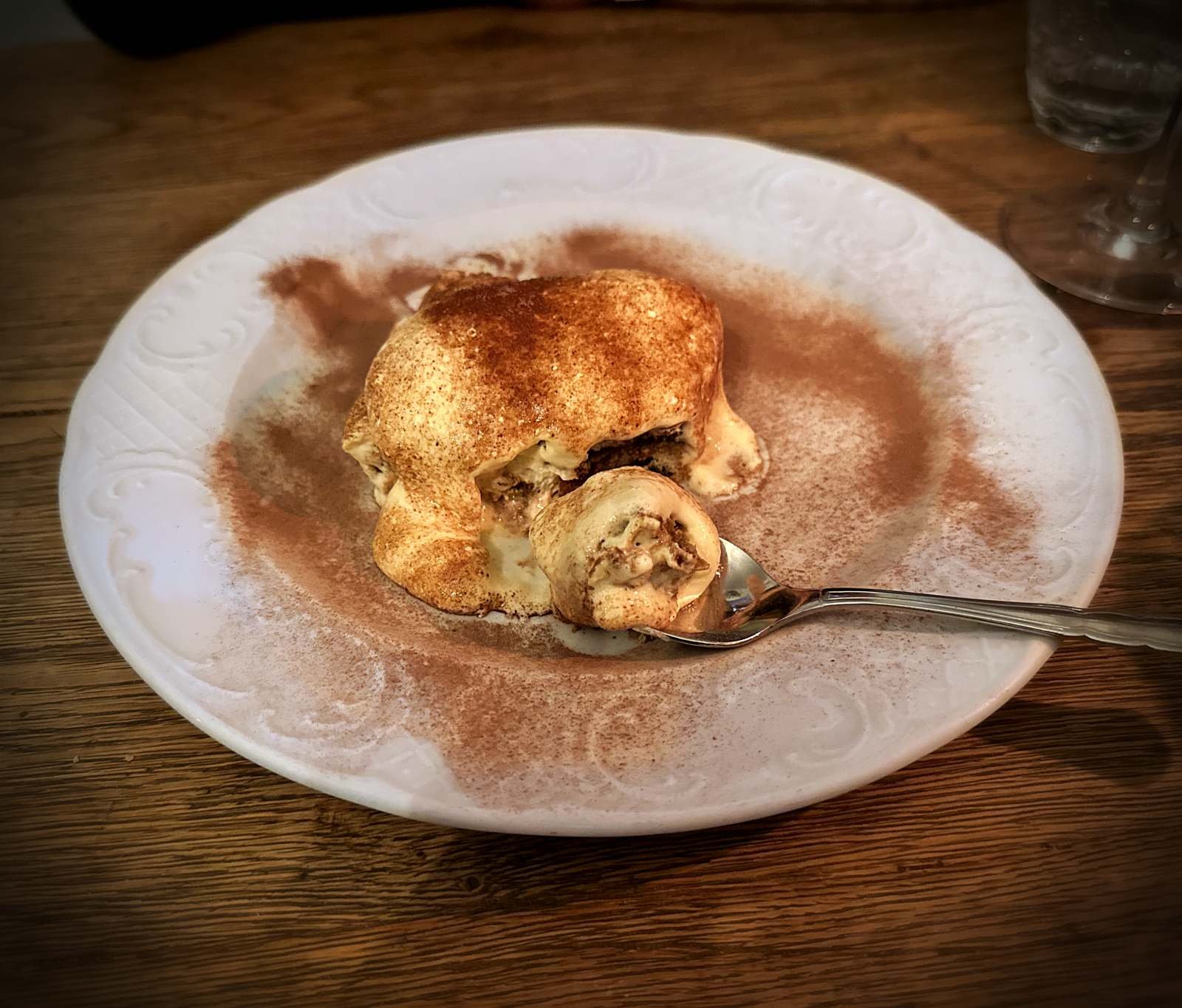 Tiramisu on a white plate dusted with cocoa powder with a spoon on a wooden table at Il Piccolo Principe restaurant in Munich.