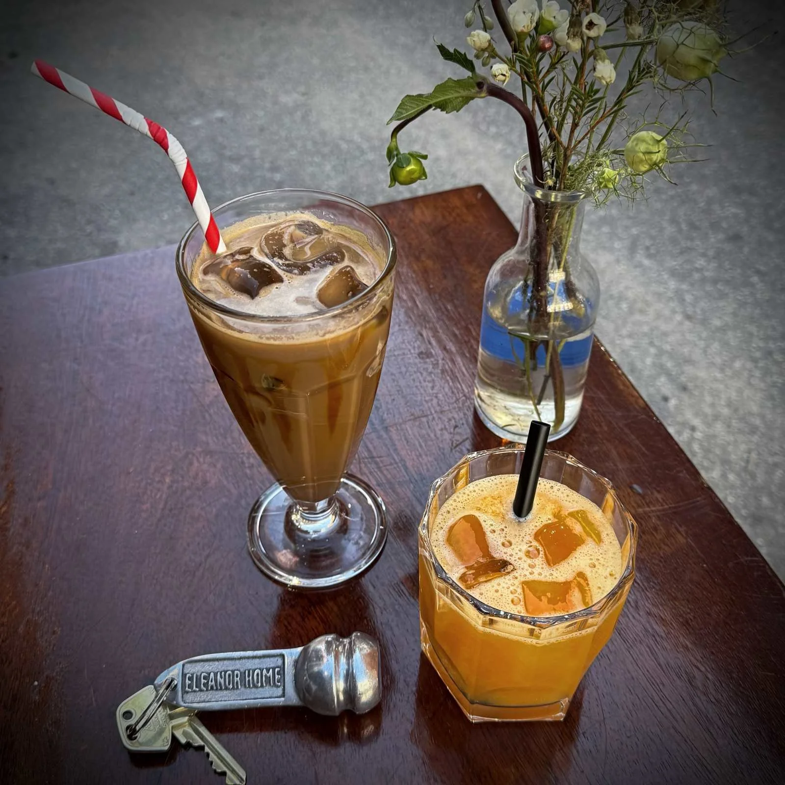 Ein Eiskaffee mit rot-weißem Strohhalm, ein orangefarbenes Getränk mit schwarzem Strohhalm, eine kleine Blumenvase und ein Zimmerschlüssel mit dem Schriftzug Eleanor Home auf einem Holztisch.