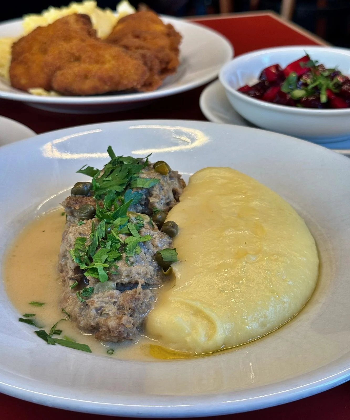 Close-up of a plate with meatballs in caper sauce and mashed potatoes on a red table. Plates with fried chicken and beetroot salad stand in the background.